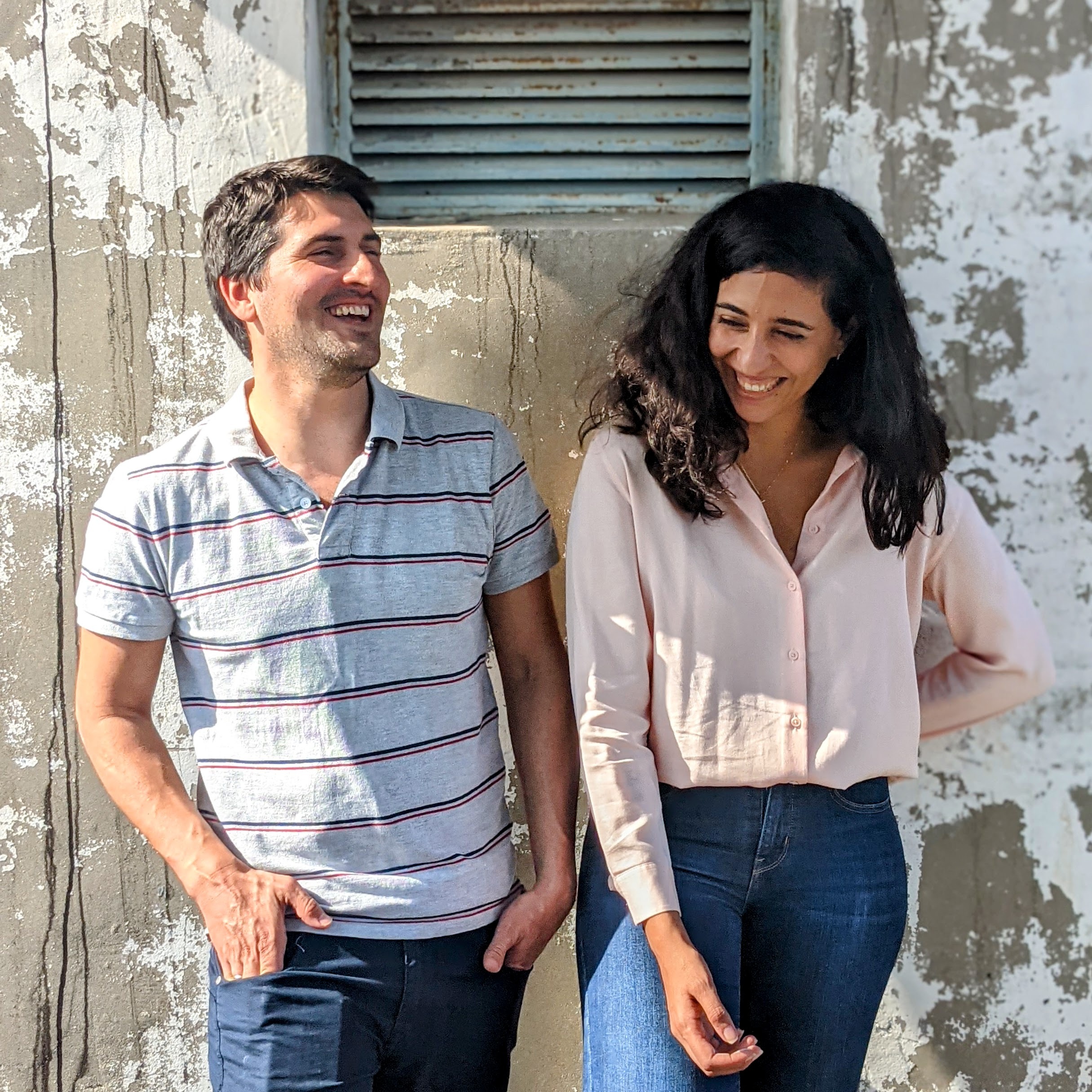A woman and a man standing side by side, their backs against a wall, and laughing