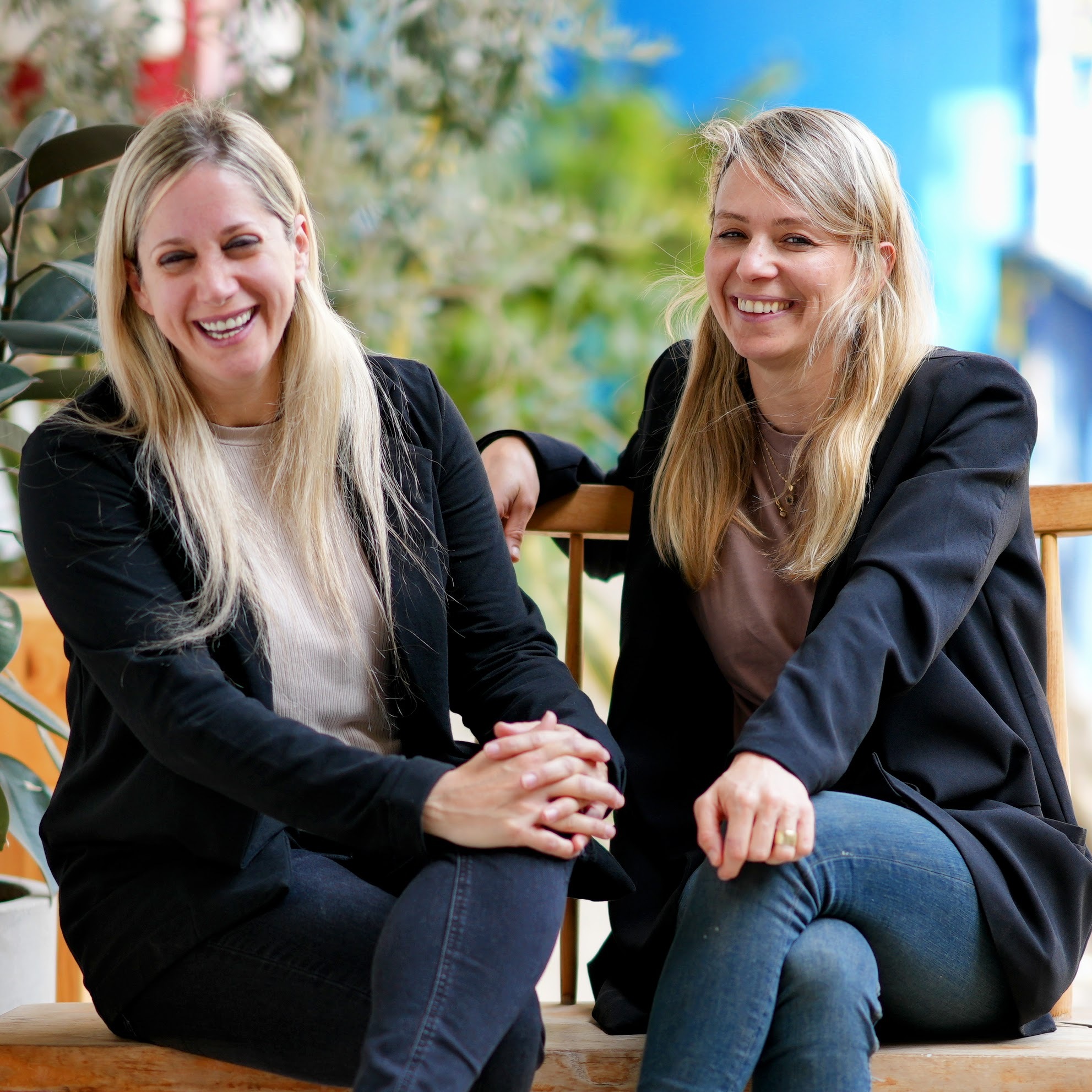 Two women sitting on a wooden bench with plants in the background, lauging